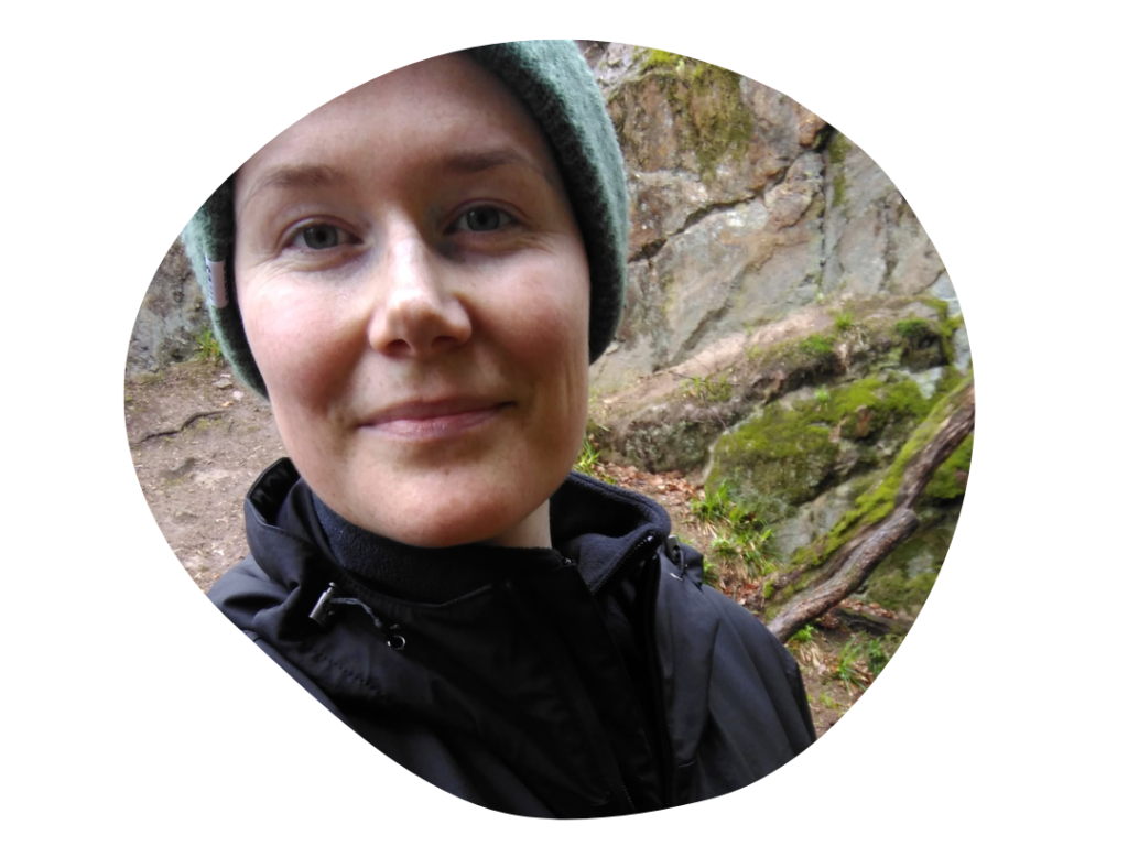 Frau Mitte 30 lächelt in die Kamera. Sie steht vor einer Felswand, die mit Moos bewachsen ist.