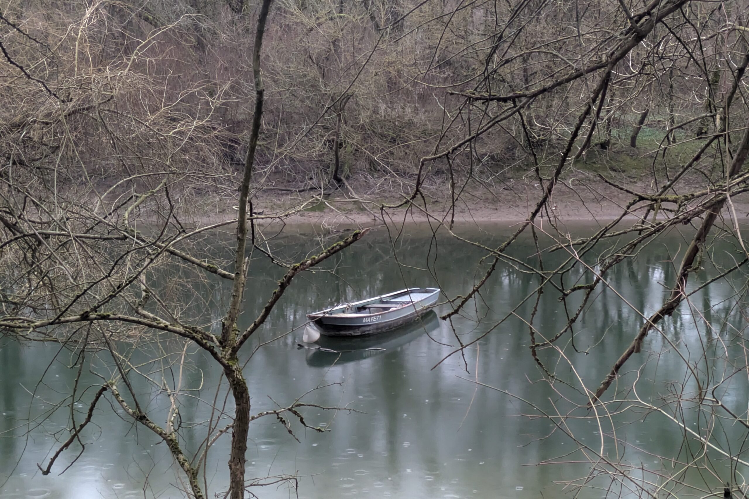 Bewaldeter Flussarm mit Regentropfen auf dem Wasser. Ein graues Boot liegt ruhig da.
