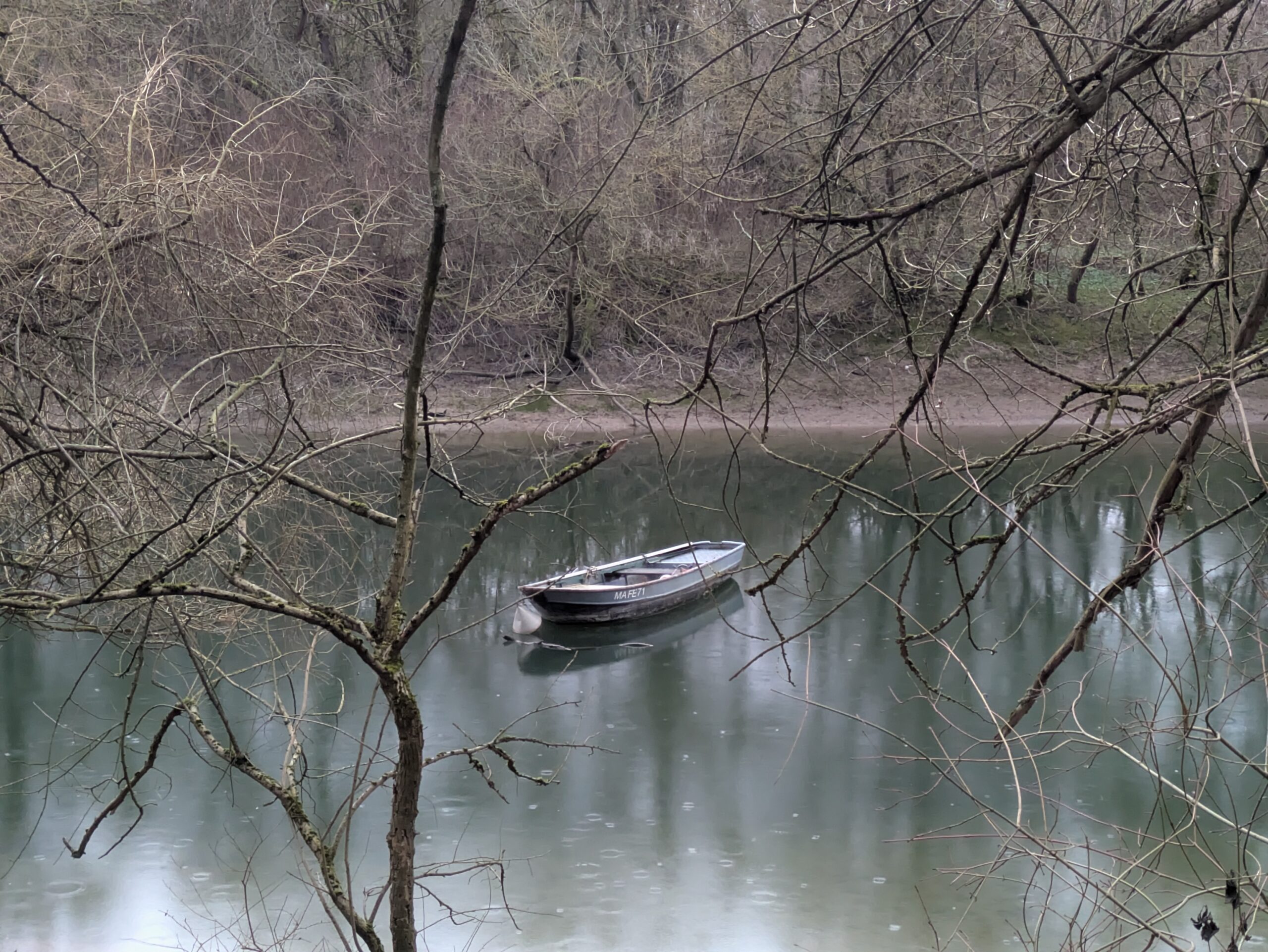 Bewaldeter Flussarm mit Regentropfen auf dem Wasser. Ein graues Boot liegt ruhig da.