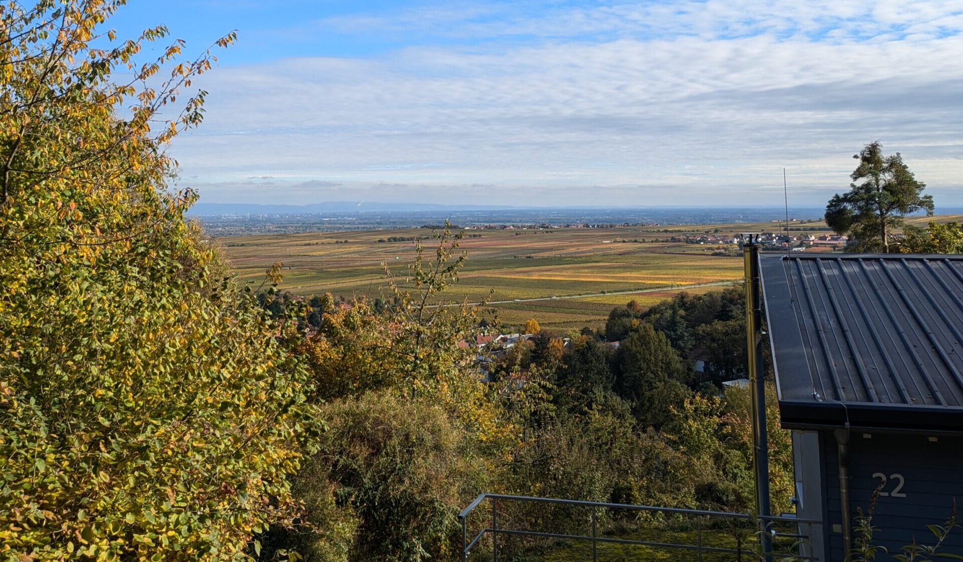 Ausblick auf Rheinebene, im Vordergrund ein Dach. Der Himmel ist blau und freundlich mit wenigen Wolken. Es ist ein sonniger Tag.